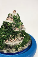 Sculpture encased in a glass tall dome with a blue clay base shaped like a turtle. Within the dome is a rocky hill landscape with moss and tiny houses.