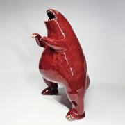 Glossy ceramic sculpture of a red stylized dinosaur with a smooth head, wide open mouth and very large body. It has tiny arms.