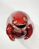 Glossy ceramic sculpture of a red stylized dinosaur with a smooth head, wide open mouth and very large body. It has tiny arms.