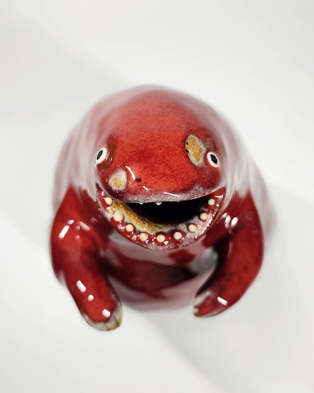 Glossy ceramic sculpture of a red stylized dinosaur with a smooth head, wide open mouth and very large body. It has tiny arms.