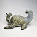 Ceramic sculpture of a sea monster, akin to Nessie the Loch Ness Monster, mostly a mossy green with a grayish blue head. It lays on its stomach and looks back over its shoulder.