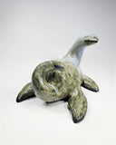 Ceramic sculpture of a sea monster, akin to Nessie the Loch Ness Monster, mostly a mossy green with a grayish blue head. It lays on its stomach and looks back over its shoulder.