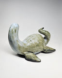 Ceramic sculpture of a sea monster, akin to Nessie the Loch Ness Monster, mostly a mossy green with a grayish blue head. It lays on its stomach and looks back over its shoulder.