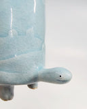 Ceramic turtle figurine with a light blue glaze and a subtle design of hills and houses on a white background