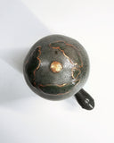 Dark grayish green ceramic turtle sculpture, with a very tall shell and a bronze line art illustration of hills and houses around its shell. Seen from overhead, where a bronze sun is at the top.