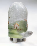 Ceramic sculpture of a turtle with a very tall shell, which has a landscape design on it of a grassy hill and small houses.