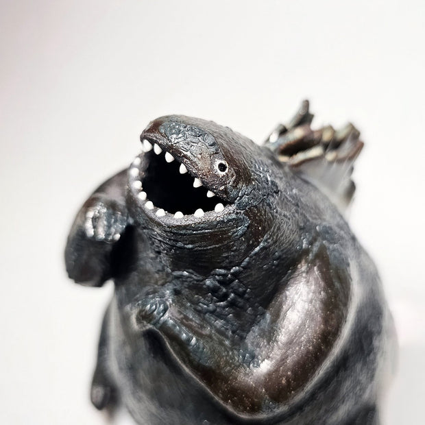 Ceramic Godzilla figure with a surface finishing akin to cooled magma, black with a subtle sheen. It has a very small, smooth head and a large plump body and back spikes.