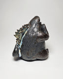 Ceramic Godzilla figure with a surface finishing akin to cooled magma, black with a subtle sheen. It has a very small, smooth head and a large plump body and back spikes.