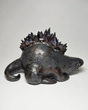 Ceramic Godzilla figure with a surface finishing akin to cooled magma, black with a subtle sheen. It has a very small, smooth head and a large plump body and back spikes. It lays on the ground