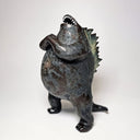 Ceramic Godzilla figure with a surface finishing akin to cooled magma, black with a subtle sheen. It has a very small, smooth head and a large plump body and back spikes. It stands and throws its head back, as if laughing.