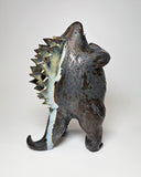 Ceramic Godzilla figure with a surface finishing akin to cooled magma, black with a subtle sheen. It has a very small, smooth head and a large plump body and back spikes. It stands and throws its head back, as if laughing.