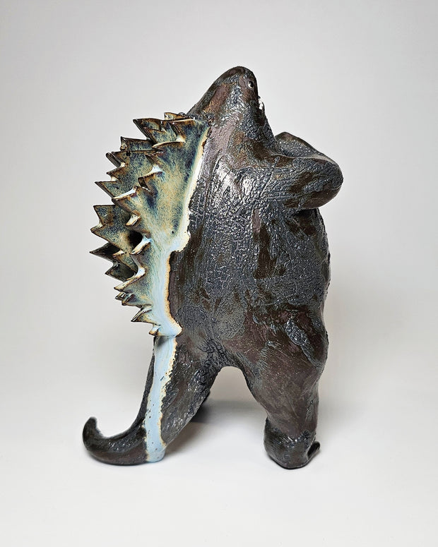 Ceramic Godzilla figure with a surface finishing akin to cooled magma, black with a subtle sheen. It has a very small, smooth head and a large plump body and back spikes. It stands and throws its head back, as if laughing.