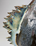 Ceramic Godzilla figure with a surface finishing akin to cooled magma, black with a subtle sheen. It has a very small, smooth head and a large plump body and back spikes. It stands and throws its head back, as if laughing.