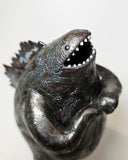 Ceramic Godzilla figure with a surface finishing akin to cooled magma, black with a subtle sheen. It has a very small, smooth head and a large plump body and back spikes. It stands and throws its head back, as if laughing.