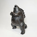 Ceramic Godzilla figure with a surface finishing akin to cooled magma, black with a subtle sheen. It has a very small, smooth head and a large plump body and back spikes. It stands and throws its head back, as if laughing.