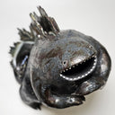 Ceramic Godzilla figure with a surface finishing akin to cooled magma, black with a subtle sheen. It has a very small, smooth head and a large plump body and back spikes. It stands and throws its head back, as if laughing.