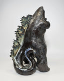 Ceramic Godzilla figure with a surface finishing akin to cooled magma, black with a subtle sheen. It has a very small, smooth head and a large plump body and back spikes. It stands and throws its head back, as if laughing.