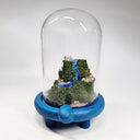 Sculpture encased in a glass tall dome with a blue clay base shaped like a turtle. Within the dome is a rocky hill landscape with moss and tiny houses and a bright blue waterfall running through it.