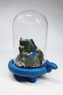 Sculpture encased in a glass tall dome with a blue clay base shaped like a turtle. Within the dome is a rocky hill landscape with moss and tiny houses and a bright blue waterfall running through it.