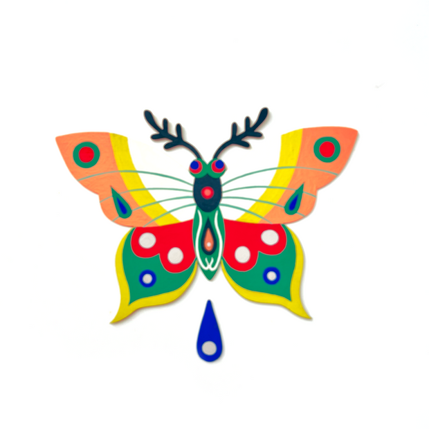 Die cut, brightly painted wooden sculpture of a moth, with textured antennae and patterned wings of many colors: white, peach, yellow, red, green and blue. A single blue water droplet hangs below the moth's body.