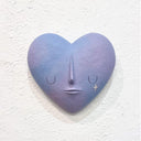 Shaped wooden sculpture of a heart, painted blue and purple with a simple closed eye expression. A single sparkle hangs from the right eye like a tear.