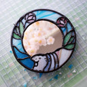 Circular mirror with a stained glass border of blue glass and purple flowers with green leaves. At the bottom is a baby hippo, light purple.