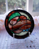 Stained glass circular hanging artwork of a brown snake made with striped glass, curled into itself and resting its head atop its body with closed eyes. Background is green and brown.