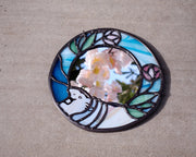 Circular mirror with a stained glass border of blue glass and purple flowers with green leaves. At the bottom is a baby hippo, light purple.