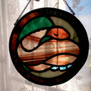 Stained glass circular hanging artwork of a brown snake made with striped glass, curled into itself and resting its head atop its body with closed eyes. Background is green and brown.