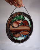 Stained glass circular hanging artwork of a brown snake made with striped glass, curled into itself and resting its head atop its body with closed eyes. Background is green and brown.