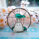 Stained glass art piece of a small white Chibi Totoro with a teal leaf overhead, surrounded by flowers.