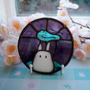 Stained glass art piece with a white rabbit and blue leaf on a checkered surface with flowers.