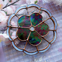 Stained glass wall hanging art of a flower, with 5 petals and a circular center. The flower is a dark, iridescent coloring and is outlined in clear, wavy glass and a blue outline.