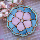 Stained glass wall hanging art of a flower, with 5 petals and a circular center. The flower is pink and is outlined in blue glass with a thinner, lighter blue outline outside of that.