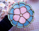 Stained glass wall hanging art of a flower, with 5 petals and a circular center. The flower is pink and is outlined in blue glass with a thinner, lighter blue outline outside of that.