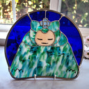 A stained glass art piece depicting a stylized character with a peach face, green and blue patterned dress, and a blue flower headpiece, set in a semicircular frame on a light surface with a window in the background.