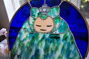 A stained glass art piece depicting a stylized character with a peach face, green and blue patterned dress, and a blue flower headpiece, set in a semicircular frame on a light surface with a window in the background.