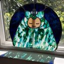 A stained glass art piece depicting a stylized character with a peach face, green and blue patterned dress, and a blue flower headpiece, set in a semicircular frame on a light surface with a window in the background.