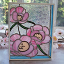 A rectangular stained glass art piece featuring three pink flowers with tiny brown dogs curled up inside of them, set in a white frame and displayed on a windowsill with blurred flowers in the foreground.