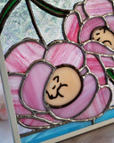 A rectangular stained glass art piece featuring three pink flowers with tiny brown dogs curled up inside of them, set in a white frame and displayed on a windowsill with blurred flowers in the foreground.