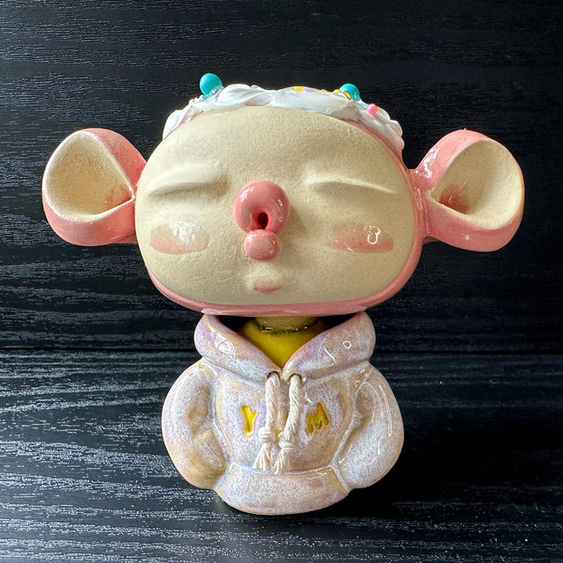 Ceramic sculpture of a large headed character, with closed eyes and full lips, making a kissy face. They have big ears and wear a hoodie, with arms tucked in. Their head is light pink with frosting atop, like a cupcake.