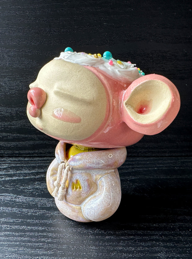 Ceramic sculpture of a large headed character, with closed eyes and full lips, making a kissy face. They have big ears and wear a hoodie, with arms tucked in. Their head is light pink with frosting atop, like a cupcake.