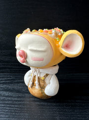 Ceramic sculpture of a large headed character, with closed eyes and full lips, making a kissy face. They have big ears and wear a hoodie, with arms tucked in. Their head is orange with sprinkles atop, like a donut.