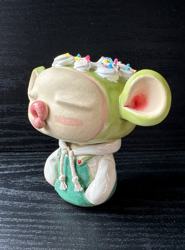 Ceramic sculpture of a large headed character, with closed eyes and full lips, making a kissy face. They have big ears and wear a hoodie, with arms tucked in. Their head is light green with frosting atop, like a cupcake.