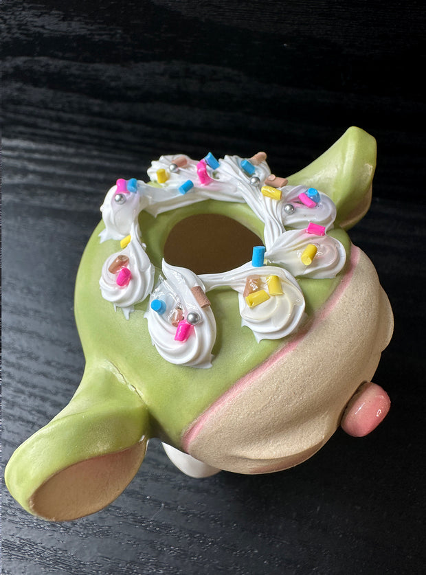 Ceramic sculpture of a large headed character, with closed eyes and full lips, making a kissy face. They have big ears and wear a hoodie, with arms tucked in. Their head is light green with frosting atop, like a cupcake.