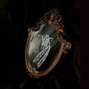 White engraved illustration on a mirror with ornate gold detailing on the outside. Image is of a fairy, with her legs pulled into her chest and an arrow through her head.