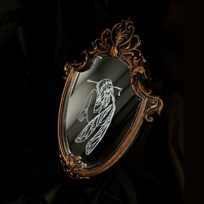 White engraved illustration on a mirror with ornate gold detailing on the outside. Image is of a fairy, with her legs pulled into her chest and an arrow through her head.