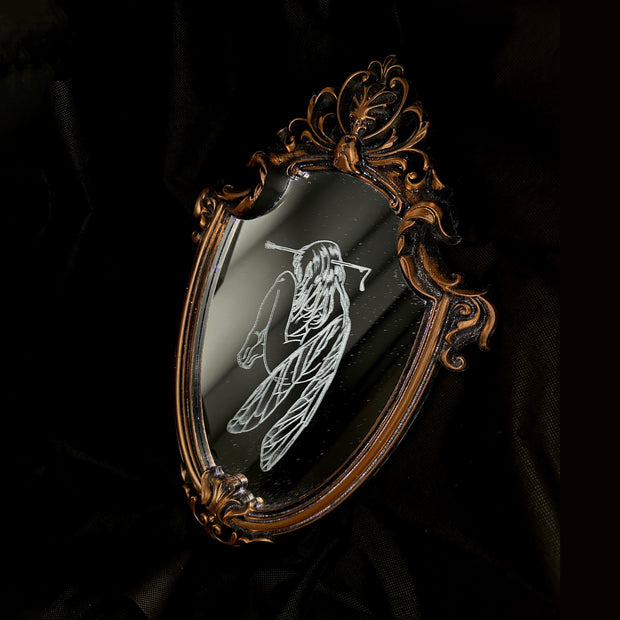 White engraved illustration on a mirror with ornate gold detailing on the outside. Image is of a fairy, with her legs pulled into her chest and an arrow through her head.