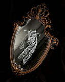 White engraved illustration on a mirror with ornate gold detailing on the outside. Image is of a fairy, with her legs pulled into her chest and an arrow through her head.