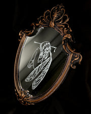 White engraved illustration on a mirror with ornate gold detailing on the outside. Image is of a fairy, with her legs pulled into her chest and an arrow through her head.